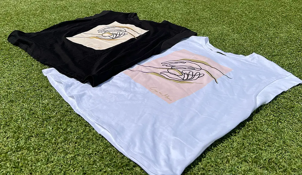 Imagen de 2 camisetas 'Coge mi mano', una blanca y otra negra, sobre un cesped