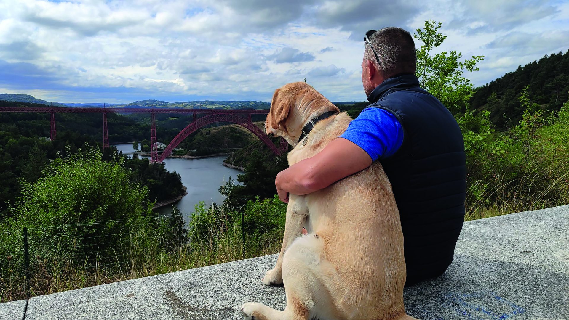 Un hombre de espaldas (David Casinos) abrazando a su perro guía, observando un río en la distancia
