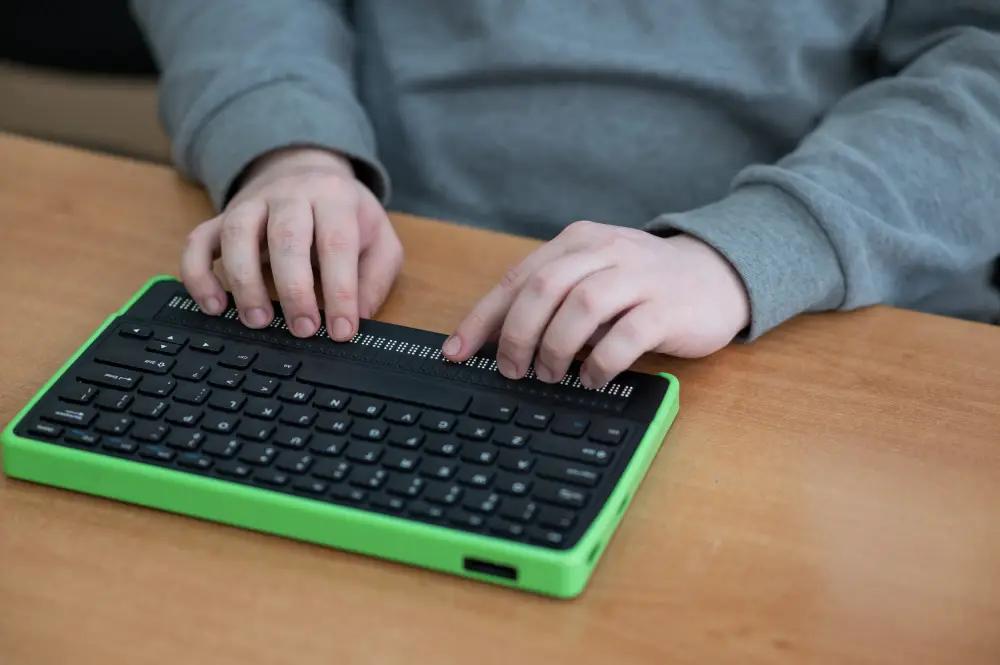 Primer plano de unas manos utilizando una línea braille integrada en un teclado adaptado para acceder a contenidos digitales.