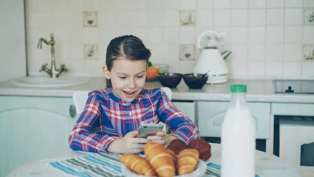 Una niña sentada a la mesa para desayunar con el móvil entre sus manos con cara de felicidad