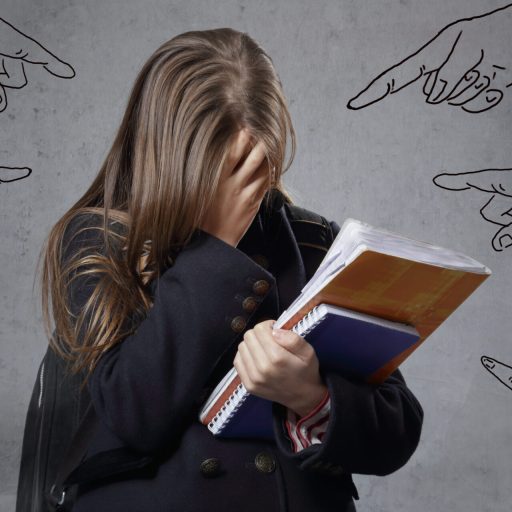 Imagen de una niña con pelo largo, sujetando libros escolares, de pie, con la mano en la cara tapándose el rostro.