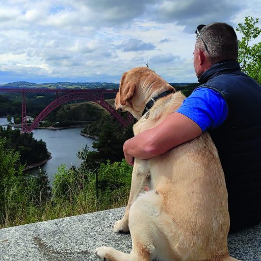 Un hombre de espaldas (David Casinos) abrazando a su perro guía, observando un río en la distancia