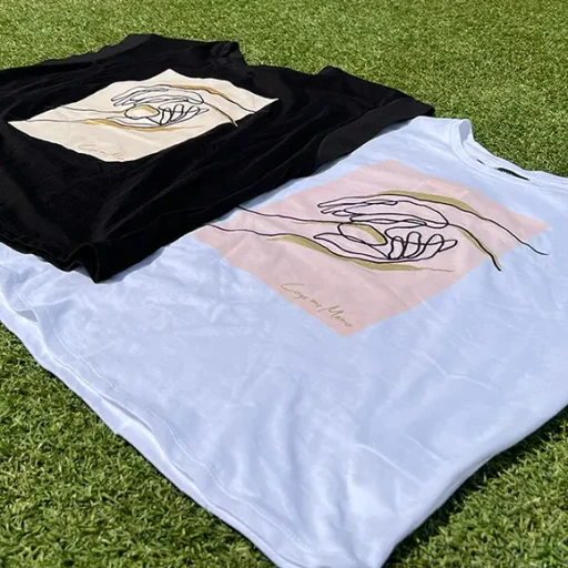 Imagen de 2 camisetas 'Coge mi mano', una blanca y otra negra, sobre un cesped